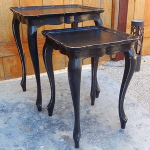May include: A set of two black nesting tables with ornate legs and a distressed finish. The tables are made of wood and have a black painted finish with gold accents.