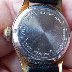 May include: Close-up of the back of a vintage wristwatch. The watch has a gold-colored bezel and a black leather strap. The watch face is silver with the words "Steel Back", "Water Resistant", and "Shock Resistant".