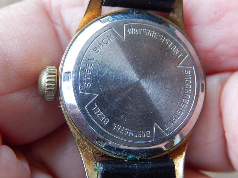 May include: Close-up of the back of a vintage wristwatch. The watch has a gold-colored bezel and a black leather strap. The watch face is silver with the words "Steel Back", "Water Resistant", and "Shock Resistant".
