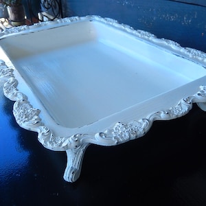 May include: A white rectangular serving tray with ornate, floral-patterned edges and small, curved feet. The tray has a distressed, vintage look, with visible brushstrokes and a slightly worn appearance. The tray is likely made of wood or a similar material.