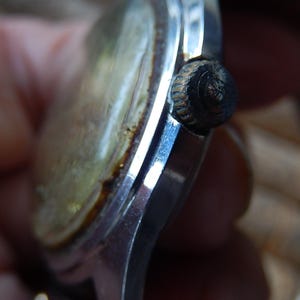 May include: Close-up of a vintage wristwatch. The watch features a silver-colored case with a tarnished, aged appearance. The watch face is a faded gold color, and the crown is black. The watch appears to be an antique.