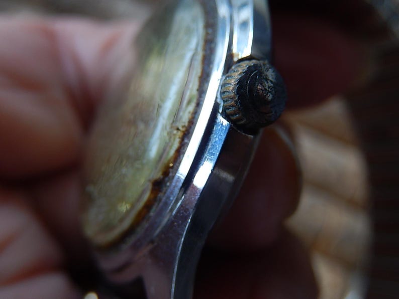 May include: Close-up of a vintage wristwatch. The watch features a silver-colored case with a tarnished, aged appearance. The watch face is a faded gold color, and the crown is black. The watch appears to be an antique.