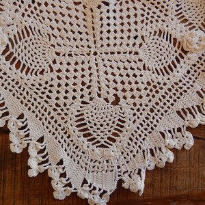 May include: A cream-colored square crochet doily featuring an intricate openwork grid pattern, central geometric motifs, and three-dimensional floral accents at the corners, finished with a decorative scalloped edge with small dangling loops.