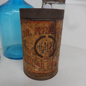 Può includere: Un'antica latta di Dr. Kraus' Health Baking Powder. Il contenitore cilindrico ha un coperchio marrone arrugginito e un'etichetta usurata con testo e un disegno a corona. Una bottiglia di vetro blu è sullo sfondo.