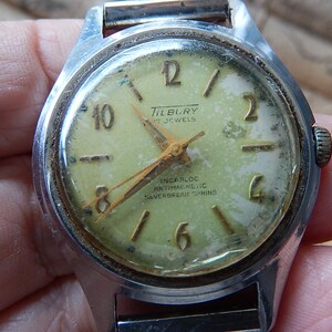 May include: Vintage Tilbury wristwatch with a silver-tone case and a faded, light green face. The watch features gold-tone hands and hour markers, with the text "Tilbury 17 Jewels" and "Incarloc Antimagnetic Neverbreak Spring".