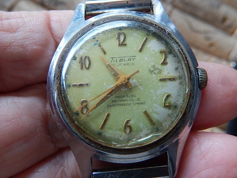 May include: Vintage Tilbury wristwatch with a silver-tone case and a faded, light green face. The watch features gold-tone hands and hour markers, with the text "Tilbury 17 Jewels" and "Incarloc Antimagnetic Neverbreak Spring".