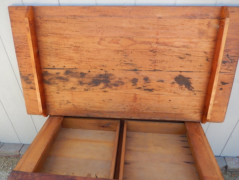 May include: A wooden table with a lift-up top and two drawers. The table is made of natural wood with a rustic finish. The top of the table is open and the drawers are empty.
