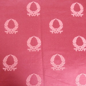 May include: A red fabric with a repeating pattern of white laurel wreaths. The wreaths are stylized and have a simple design.