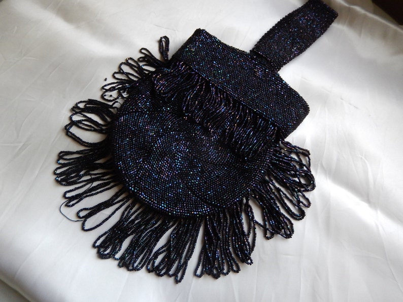 May include: A black beaded evening bag with a fringe detail. The bag is made of small, iridescent beads and has a long, beaded strap.
