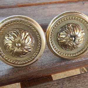 May include: Two antique brass door knobs. Each knob features a detailed floral design in the center, surrounded by a beaded border. The knobs are round and have a warm, golden hue, suggesting they are made of brass.