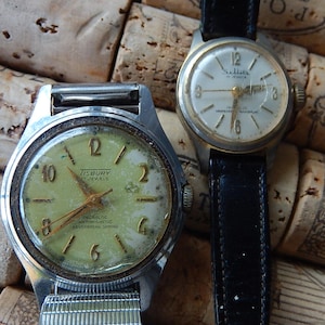 May include: Two vintage wristwatches with aged faces and metal cases. One watch has a light green face with the text "TISBURY 17 JEWELS". The other watch has a white face and a black leather band.