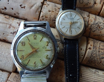 Op de afbeelding: Twee vintage polshorloges. Eén heeft een lichtgroene wijzerplaat met de tekst "TISBURY 17 JEWELS". De andere heeft een witte wijzerplaat en een zwarte leren band. Beide horloges hebben metalen kasten en verouderde wijzerplaten.
