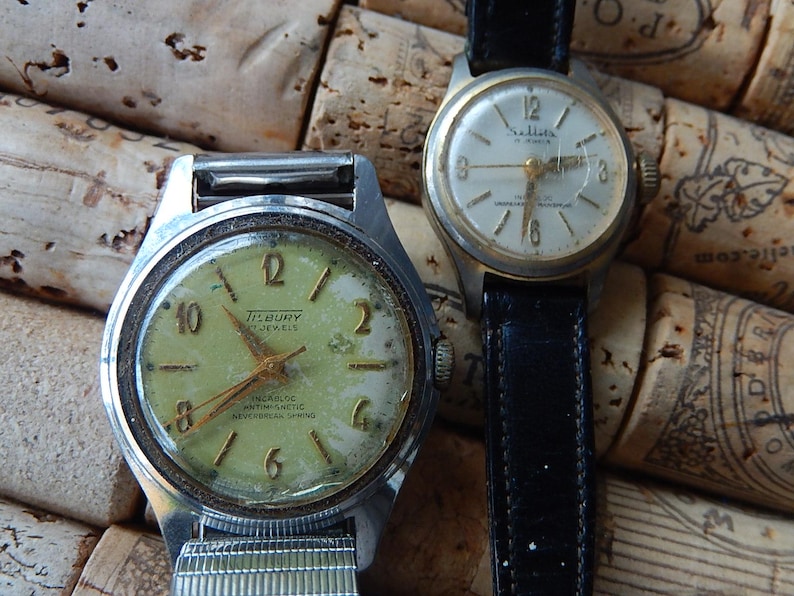 May include: Two vintage wristwatches with aged faces and metal cases. One watch has a light green face with the text "TISBURY 17 JEWELS". The other watch has a white face and a black leather band.