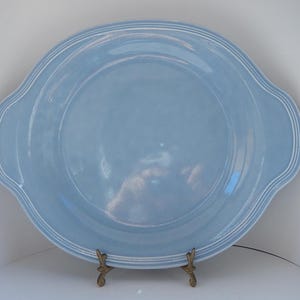 May include: A large, round, light blue ceramic serving platter with two handles. The rim has a subtle white trim. The platter is displayed on a decorative bronze stand. This dish is suitable for serving food.