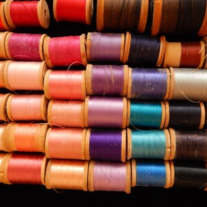 Pu&ograve; includere: Una collezione di bobine di filo di vari colori, tra cui rosso, rosa, viola, blu, verde e giallo. Le bobine sono disposte in file, con il filo avvolto attorno a bobine di legno.
