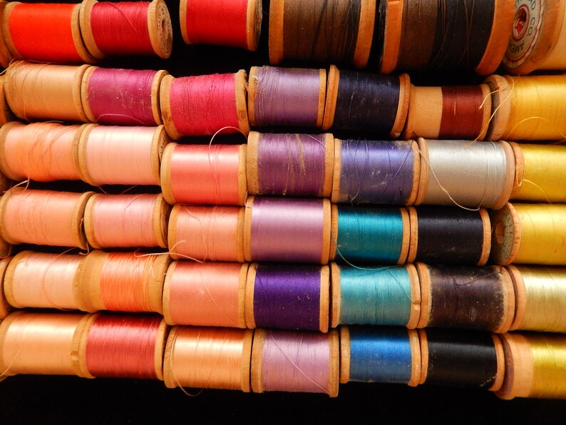 Pu&ograve; includere: Una collezione di bobine di filo di vari colori, tra cui rosso, rosa, viola, blu, verde e giallo. Le bobine sono disposte in file, con il filo avvolto attorno a bobine di legno.
