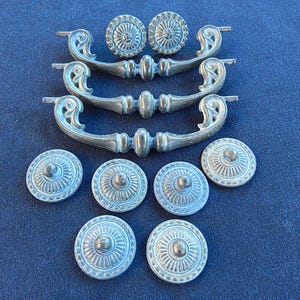 May include: A collection of three silver-toned metal drawer pulls with curved, ornate handles and six matching circular knobs featuring a radial, sunburst-like pattern. The hardware has a weathered, matte finish with visible detailing.