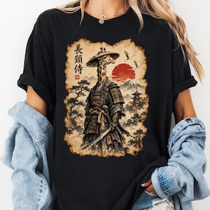 May include: A black t-shirt featuring a graphic print of a giraffe dressed as a samurai warrior. The giraffe wears traditional armour and a conical hat, holding a katana. The background shows a red sun, a mountain, trees, and Japanese text.