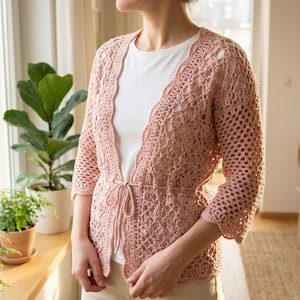 Peut inclure: Un gilet en crochet rose clair avec un motif ajouré, des manches trois-quarts et un lien de serrage fin à la taille. Le vêtement est porté sur un t-shirt blanc uni et un pantalon clair, mettant en valeur un bord festonné.