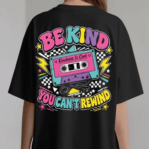 Può includere: Una t-shirt nera con una grafica colorata sul retro. Il design presenta una cassetta audio con il testo "Kindness Is Cool" e le frasi "BE KIND" e "YOU CAN'T REWIND" in caratteri audaci e multicolori, arricchite da stelle e fulmini.