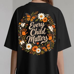 Puede incluir: Una camiseta negra con un gráfico circular en la parte posterior. El diseño incluye el texto 'Every Child Matters' en una fuente cursiva color crema, rodeado de mariposas monarca naranjas, margaritas blancas, flores naranjas, hojas verdes, dos plumas y dos huellas de manos naranjas.