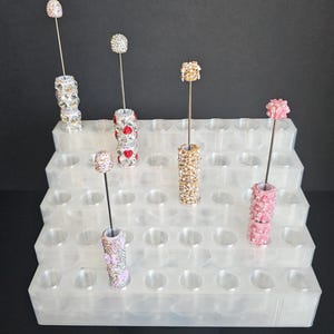 May include: An assortment of decorative hat pins with jewel-encrusted cylindrical bases and spherical or floral tops. The pins are displayed in a tiered, clear plastic organizer with multiple circular holes. The pins feature silver, gold, red, and pink accents.