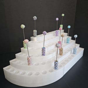 May include: A tiered white display stand holds several decorative floral arrangements. Each arrangement features a beaded cylinder base and a spherical, colorful beaded flower. The stand has multiple levels with circular holes to hold the arrangements.