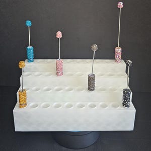 May include: A white tiered display stand holding several decorative items. Each item has a cylindrical base covered in colorful rhinestones, topped with a rhinestone-covered sphere. The stand has multiple holes to hold the items upright.