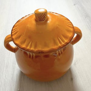 Puede incluir: Una olla de cerámica de color naranja brillante con una tapa a juego y dos asas laterales. La tapa presenta un borde festoneado texturizado y un pequeño pomo con ranuras verticales en el centro. La olla tiene un cuerpo redondeado y liso.