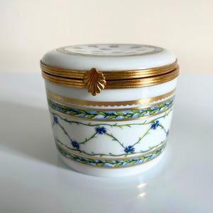 Può includere: Un portagioie in porcellana bianca con bordo e chiusura in metallo color oro. La scatola presenta un motivo floreale con fiori blu e foglie verdi. Il coperchio ha un motivo di piccoli fiori blu.