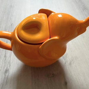 Op de afbeelding: Een feloranje keramische theepot in de vorm van een olifant. De theepot heeft een gebogen handvat aan de linkerkant, een deksel met een kleine knop aan de bovenkant en een olifantenkop met een slurf als tuit en grote oren aan de zijkanten.