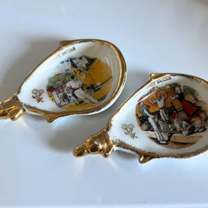 Peut inclure: Deux cendriers vintage en porcelaine blanche avec des bordures dorées et une forme unique en forme de dauphin. Chaque cendrier présente une illustration colorée de personnes en costume traditionnel, avec le texte "PAYS BASQUE" en lettres dorées.