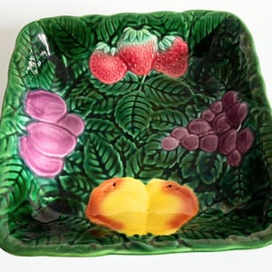 Könnte beinhalten: Ein quadratischer, grüner Keramikservierteller mit einem erhabenen Blättermuster. Der Teller ist mit Erdbeeren, Trauben, Pfirsichen und rosa Blumen verziert. Die Früchte und Blumen sind in leuchtenden Farben gehalten.