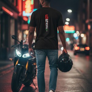 May include: A black t-shirt with the text "65432N1" in red and green, worn by a person walking away from the camera. The person is holding a black helmet and is near a black motorcycle. The background is a city street at night.