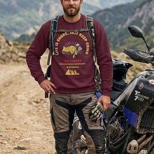 May include: A maroon sweatshirt with the text "LIFE IS SHORT, JUST KEEP GOING" and a graphic. The person is wearing hiking pants, boots, and a backpack, standing next to a motorcycle. The motorcycle is black and blue.