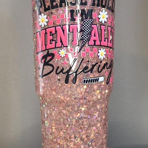 May include: A clear, glitter-filled tumbler with the text "PLEASE HOLD I'M MENTALLY BUFFERING" in pink and black. The tumbler features daisy accents and a lightning bolt graphic. The cup is displayed on a white stand.