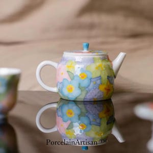 Può includere: Una piccola teiera in ceramica dipinta a mano con un motivo floreale in colori pastello. La teiera presenta un manico, un beccuccio e un coperchio bianchi con un pomello blu. Il design include fiori blu, gialli e viola.