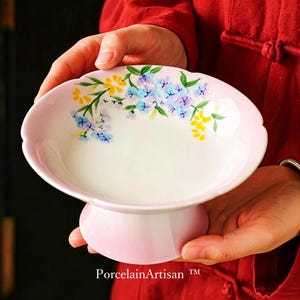 Op de afbeelding: Een roze porseleinen voetbakschaal met een geschulpte rand, versierd met handgeschilderde blauwe, gele en groene bloemaccenten. De schaal wordt voor een rode achtergrond gehouden. De tekst "PorcelainArtisan" is onderaan zichtbaar.