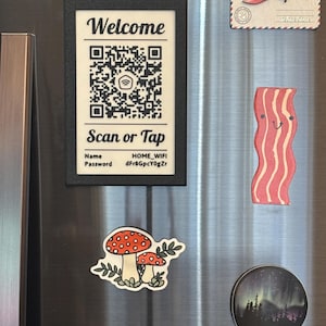 May include: A black framed sign with the word "Welcome" above a QR code and the words "Scan or Tap". Also, a red airplane magnet, a bacon magnet, a mushroom magnet, and a round magnet with a forest scene.