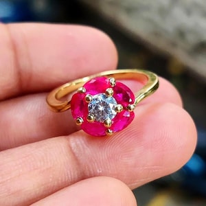 Puede incluir: Un anillo de oro con un diseño en forma de flor. El anillo tiene una piedra central clara y redonda, rodeada de gemas de color rosa vibrante. Pequeños detalles dorados se colocan alrededor de las piedras rosas, realzando el diseño general del anillo.