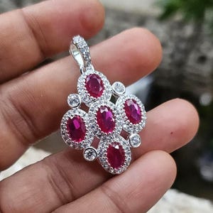 May include: A silver-toned pendant featuring five oval-cut deep red stones arranged in a cluster. Each red stone is surrounded by a halo of small, clear, round-cut stones. The pendant includes a silver-toned bail at the top for attachment.