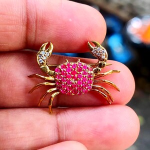 Puede incluir: Broche de cangrejo dorado con un cuerpo incrustado con pequeñas piedras preciosas de color rosa brillante. Las pinzas del cangrejo están adornadas con piedras claras y brillantes. La broche tiene un alfiler en la parte posterior.