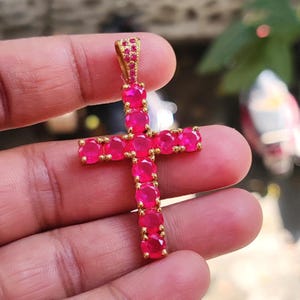 Könnte beinhalten: Ein goldfarbener Anhänger in Form eines Kreuzes, verziert mit leuchtend rosa Edelsteinen. Die Edelsteine sind in einen goldfarbenen Metallrahmen gefasst, was einen auffälligen Kontrast erzeugt. Der Anhänger hat oben eine Schlaufe für eine Kette.