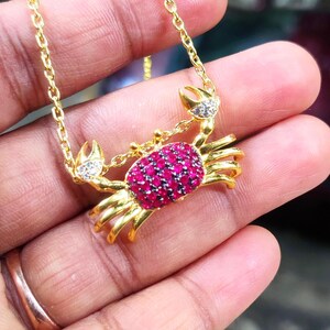 May include: A gold-toned necklace featuring a crab pendant. The crab's body is encrusted with numerous small, vibrant pink stones, while its two raised claws are accented with small, clear, sparkling stones. The pendant hangs from a gold chain.