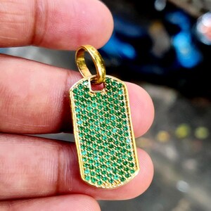 Puede incluir: Colgante dorado en forma de placa rectangular, incrustado con numerosas piedras preciosas verdes pequeñas. El colgante tiene un lazo dorado en la parte superior para una cadena. Las piedras preciosas están muy juntas, creando una superficie vibrante y texturizada.