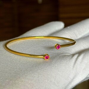 Puede incluir: Una pulsera abierta de tono dorado con una banda lisa y pulida. Los dos extremos de la pulsera terminan en pequeños engastes circulares, cada uno con una piedra preciosa rosa de corte redondo y vibrante orientada hacia adentro.