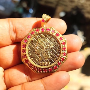 Könnte beinhalten: Goldener Anhänger mit Münzdesign, das das Profil einer Frau und das Jahr 1886 zeigt. Der achteckige Rahmen ist mit kleinen roten Edelsteinen verziert. Der Anhänger hat eine Schlaufe für eine Kette.