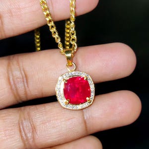 May include: A gold-toned necklace featuring a square-shaped pendant with a vibrant red cushion-cut center stone. The pendant is framed by a border of small, clear, sparkling stones and is suspended from a matching gold-toned curb chain link.