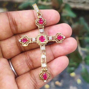 Può includere: Un pendente a forma di croce color oro con pietre preziose rosse e piccole pietre trasparenti. La croce presenta un design dettagliato con una pietra rossa centrale e ulteriori pietre rosse alle estremità delle braccia della croce. Il pendente è tenuto da un piccolo anello.