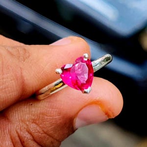 May include: A silver-toned ring featuring a prominent heart-shaped, vibrant pink gemstone set in a prong setting. The band is smooth and metallic, showcasing a polished finish that reflects light, highlighting the faceted cut of the central stone.
